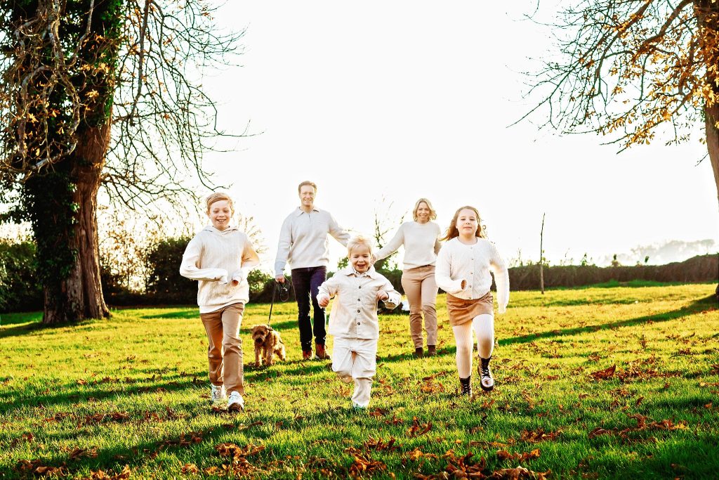 A joyful happy family with 3 young children and a dog run on the grass towards the viewer
